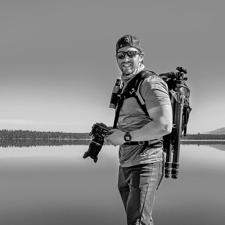 Artist Greg Piazza with camera equipment standing on a rock by a lake